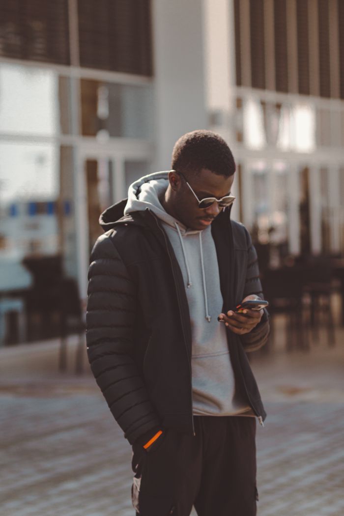 Contact Young man in a hoodie and sunglasses checks his smartphone outside a modern building.
