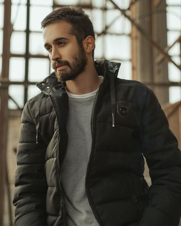 Home A thoughtful bearded man wearing a black jacket poses indoors with soft lighting.