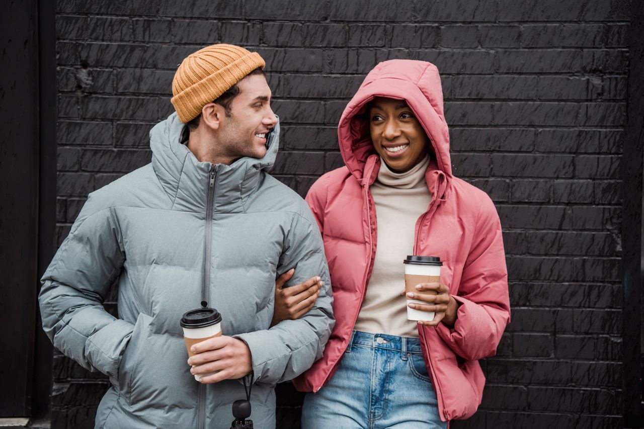 Services Content diverse couple in casual outerwear with paper cups of tasty takeaway coffee while standing in city on cold day
